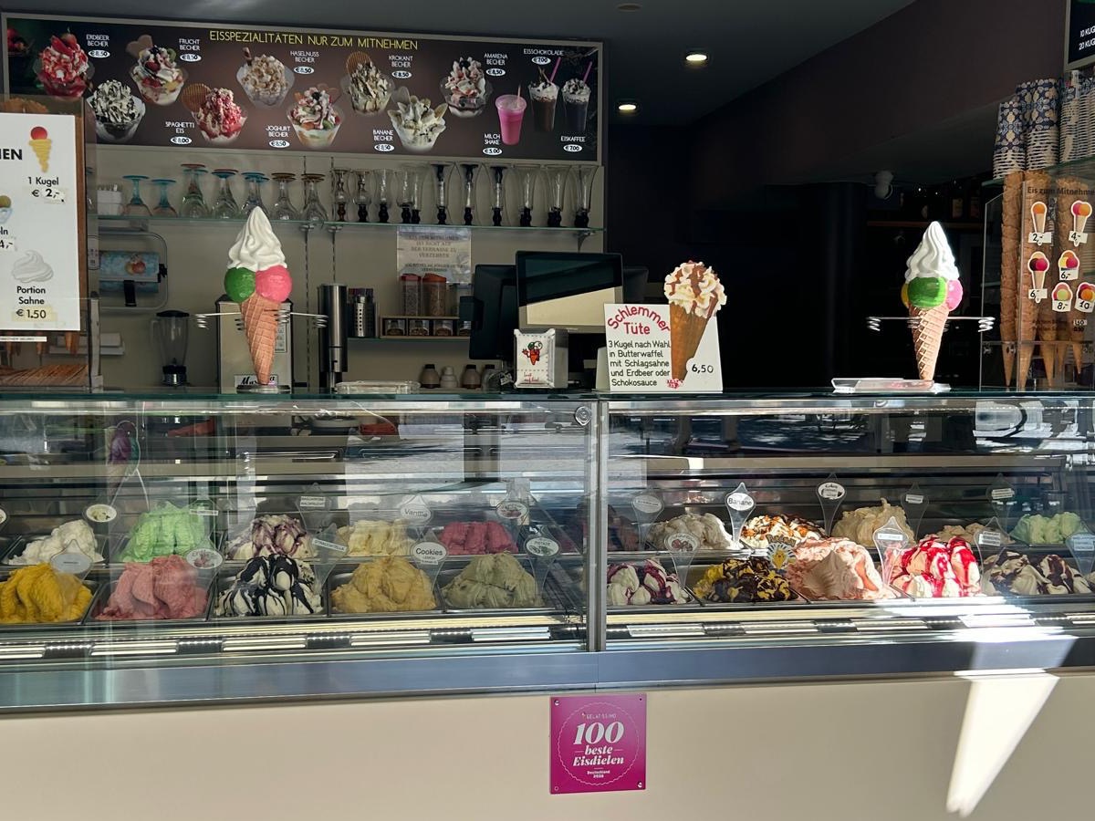 Die Eisvitrine des Capri: rund zwanzig Sorten hausgemachtes Gelato in offenen Behältern, darüber die Tafel mit Eisbechern und der Plakette „100 beste Eisdielen“