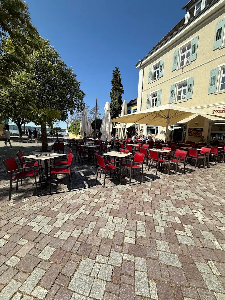 Die Terrasse des Capri am Landungsplatz in Überlingen: rote Stühle und Sonnenschirme in der Mittagssonne, hinter den Bäumen der Bodensee